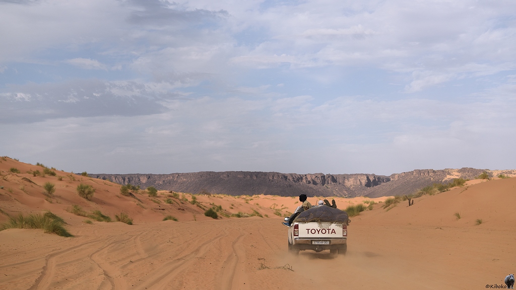 Das Bild zeigt einen vorausfahrenden weißen Geländewagen mit zwei Personen auf der Ladefläche. Der Wagen fährt durch losen Sand in ein enges Tal. Im Hintergrund ist ein breites Tal, welches durch eine Bergkette aus dunklen Felsen begrenzt wird.