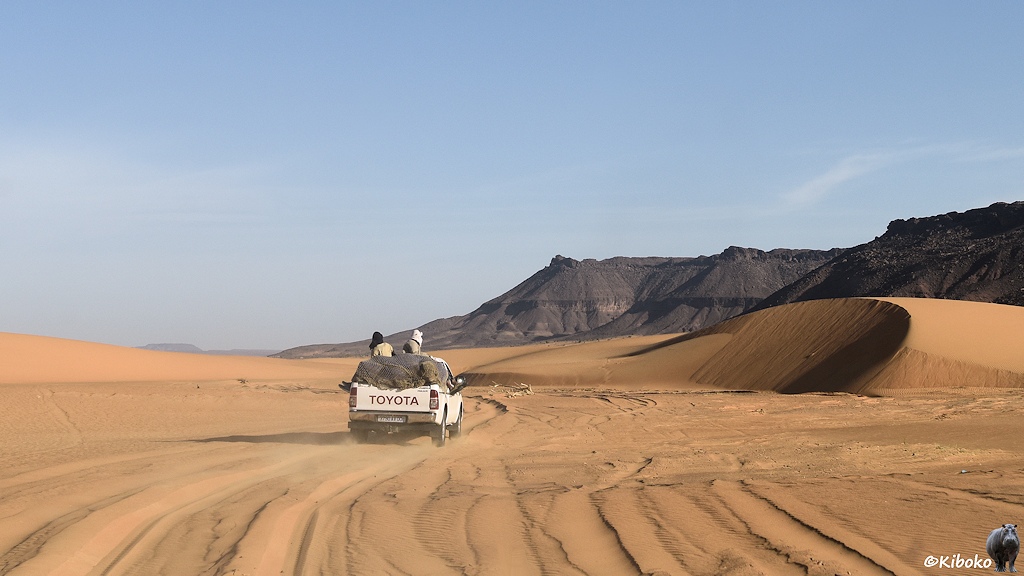 Das Bild zeigt einen vorausfahrenden weißen Geländewagen, auf dem zwei Männer auf der Ladfläche sitzen. Der Wagen fährt durch hellbraunen Tiefsand in einer tiefen Spur. Auf beiden Seiten sind Dünen.