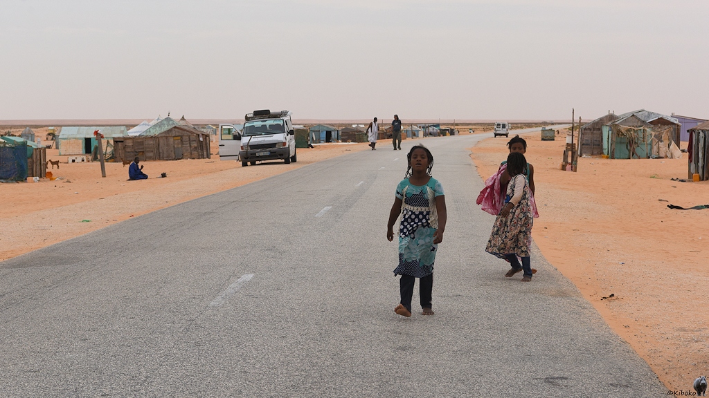 Das Bild zeigt eine Teerstraße durch einen Ort aus einfachen Hütten. Drei Mädchen in bunten Kleidern spielen auf der Straße. Am linken Straßenrand steht ein weißer Kleinbus mit geöffneter Beifahrertür. Vor dem Auto kniet ein Mann im Sand und betet Richtung Osten - nach Mekka.