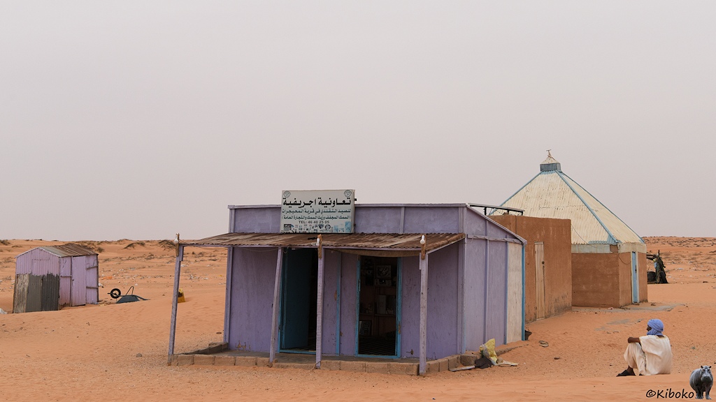 Das Bild zeigt ein lilafarbenes Gebäude mit zwei Türen und einem Vordach. Darüber ist ein Schild in arabischer Schrift. Ein Mann mit weißem Gewand und hellblauem Turban sitzt rechts neben dem Haus. Links ist eine kleine Hütte in der selben Farbe. Dahinter sind weitere Gebäude. Ein Haus hat ein hohes Walmdach aus Blech.