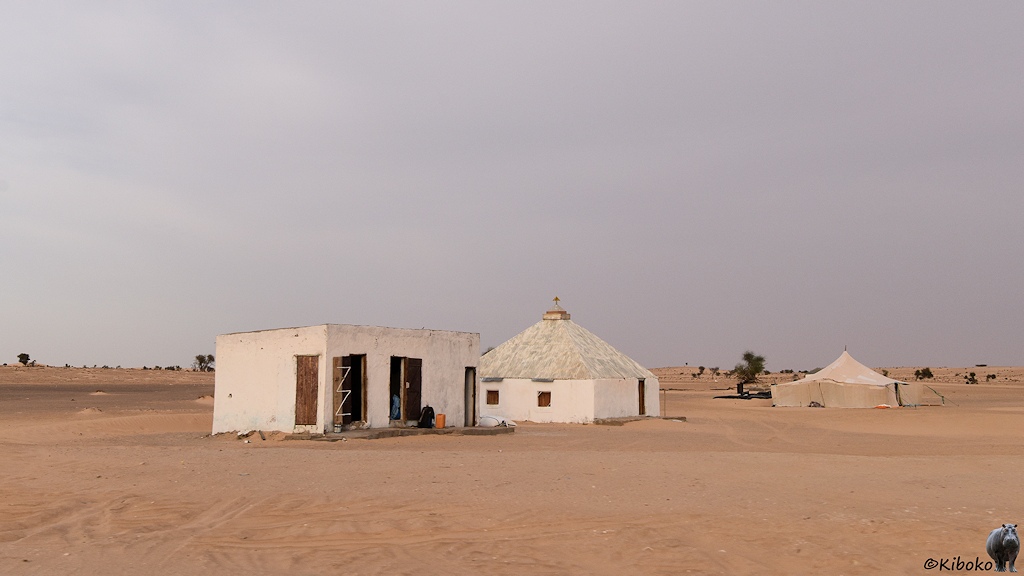 Das Bild zeigt ein kleines weißes Haus mit Flachdach. dahinter ist ein quadratisches Haus mit einem Walsmdach aus Wellblech. Recht daneben steht ein weißes, quadratisches Zelt mit einer hohen Mittelstütze. Dahinter ist nur hellbraune Sandwüste.