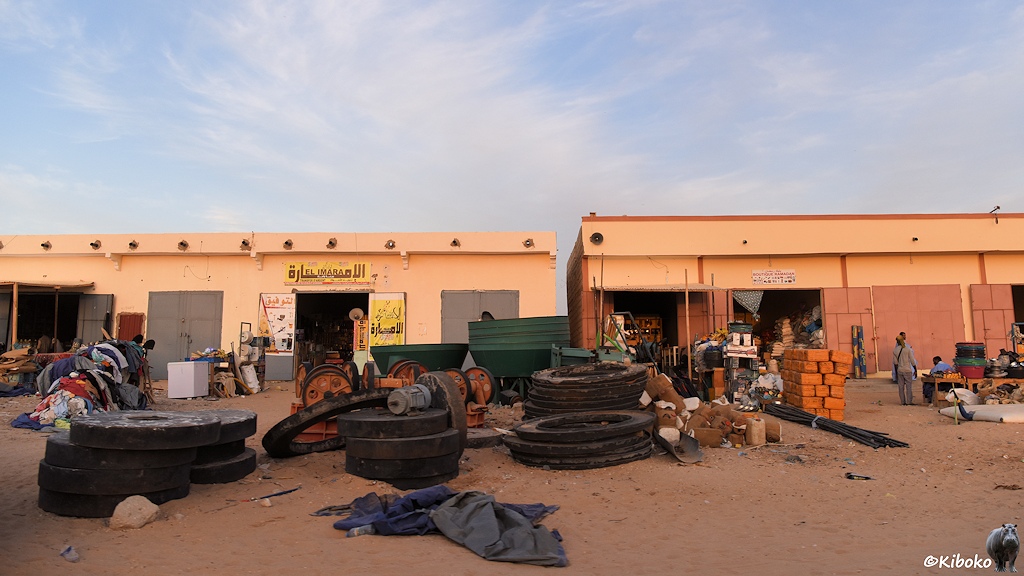 Das Bild zeigt zwei weitere Geschäftshäuser. Vor den Toren sind große Steinscheiben, Trichter, Kanister, Armierungsstahl, Sand und Müll.