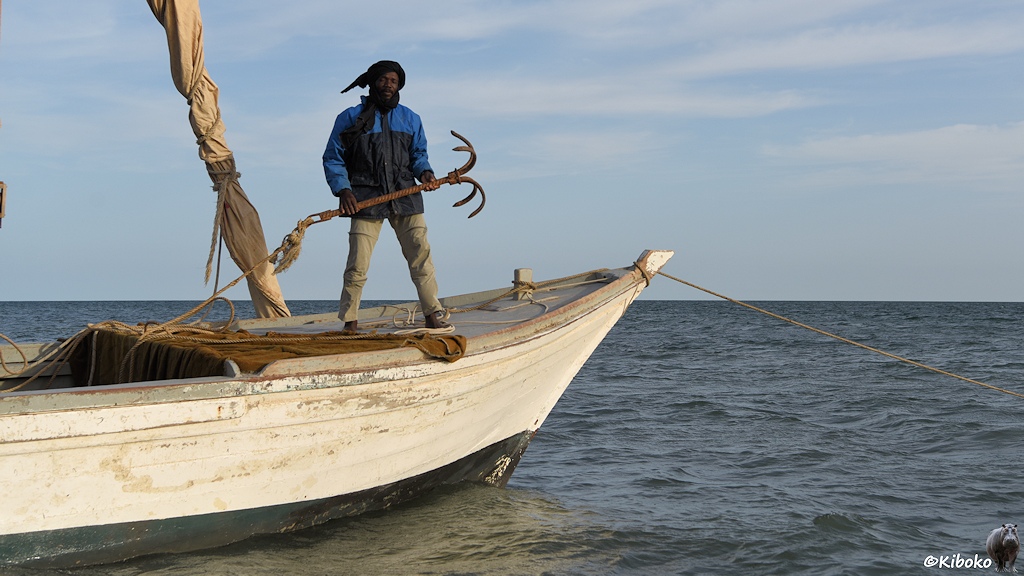 Das Bild zeigt einen Mann in blauer Jacke und grauer Hose, der mit eiden Händen einen Anker aus gebogenen Armierungseisen hält. Er steht auf der Spitze eines weißen Segelbootes aus Holz. Das gerefte Segel steht link neben ihm.