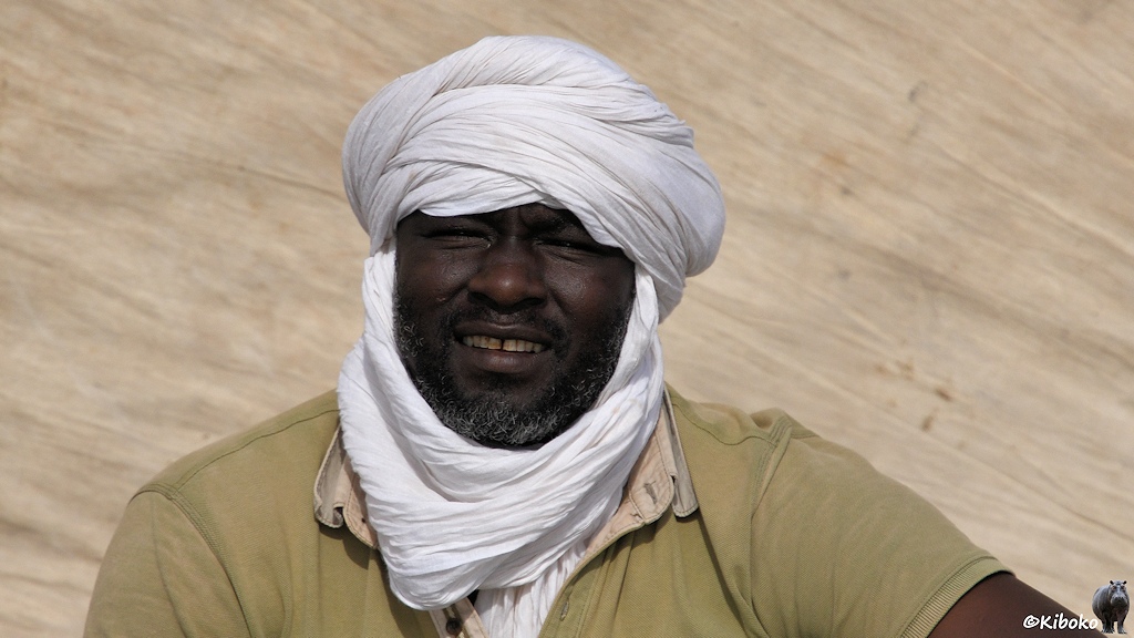 Das Bild zeigt einen Mann mit Vollbart in beigefarbenen T-Shirt mit weißem Turban vor einem beigefarbenen Segel. Er lächelt in die Kamera.