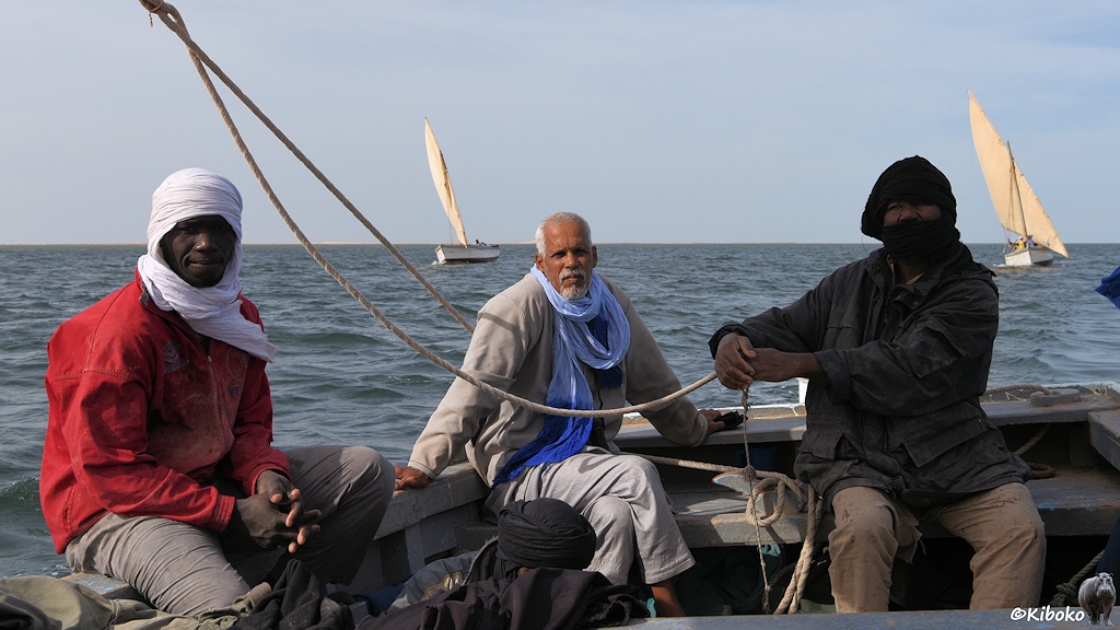 Das Bild zeigt drei Männer im Heck eines Segelbootes. Der rechte Mann mit dunkler Jacke und schwarzem Turban hält das Seil für das Segel fest. In der Mitte sitzt ein älterer Mann mit weißem Bart. Den hellblauen Turban hat er für das Foto abgelegt. Links sitzt ein weiterer Mann in roter Jacke und weißem Turban auf der Bordwand. Im Hintergrund sind zwei weitere Segelboote.