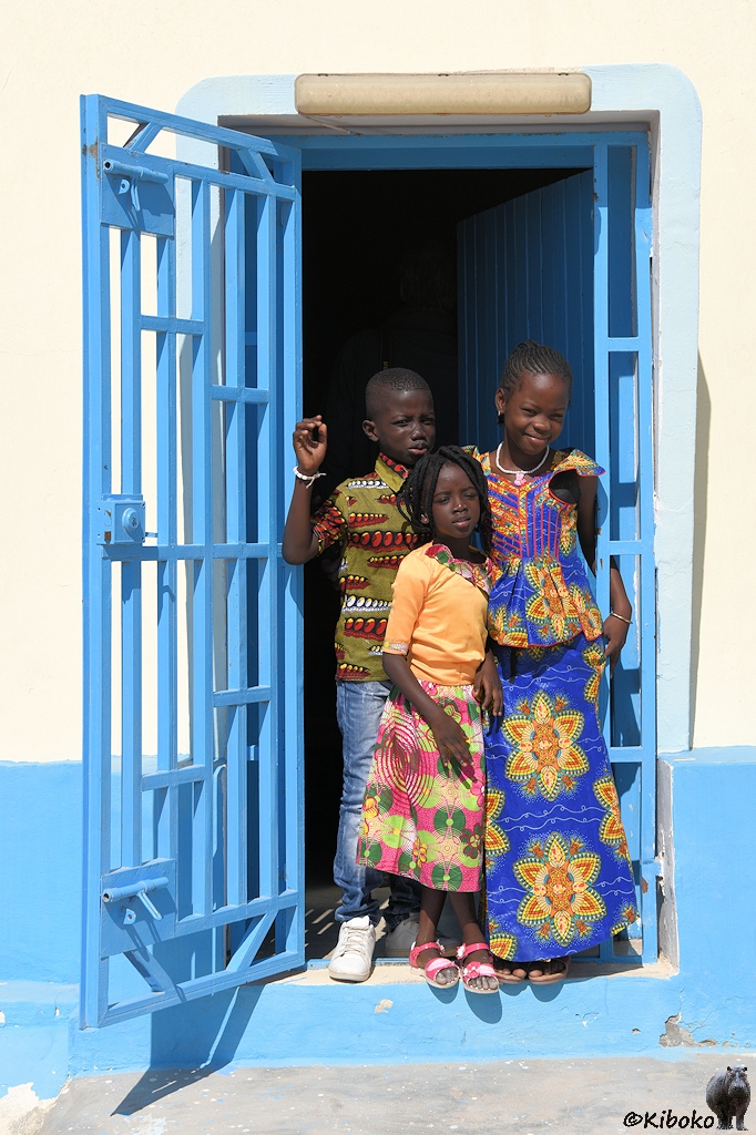 Das Bild zeigt eine Hochformataufnahme einer offenen, hellblauen Tür. Ein Metallgittertor ist außen nach links geöffnet. Eine Holztür ist innen nach rechts geöffnet. In der Tür stehen drei Kinder, zwei Mädchen und ein Junge. Sie tragen bunte Kleider in afrikanischen Mustern.