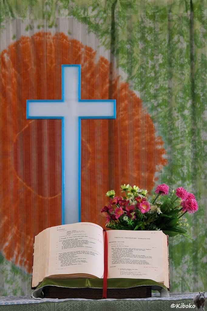 Das Bild zeigt eine Hochformataufnahme des weißen Kreuzes auf dem Altar. Das Kreuz hat eine hellblaue Umrandung. Daneben ist ein Strauß mit bunten Blumen. Vor dem Kreuz ist eine aufgeschlagene Bibel. Hinter dem Kreuz ist ein Vorhang mit einer großen orangeroten Fläche hinter dem Kreuz.
