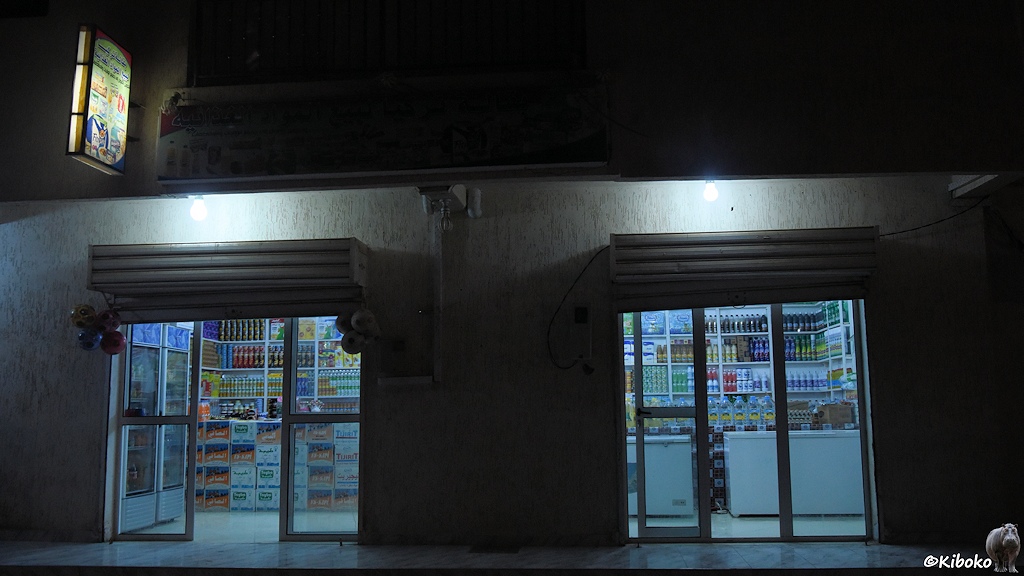 Das Bild zeigt eine Nachtaufnahme von einem Supermarkt. Zwei doppelte Glastüren erlauben einen Blick in den beleuchteten Laden. In den Wandregalen stehen Plastikflaschen mit Getränken. Davor stehen Pappkartonstapel und weiße Kühltruhen.
