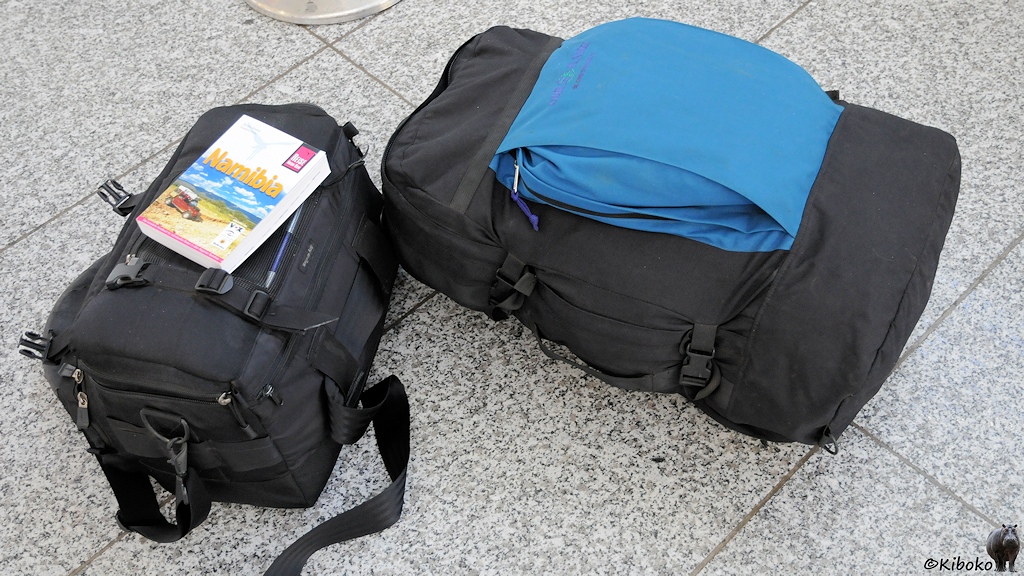 Auf dem Boden im Terminal liegt ein dunkelblauer Rucksack, eine schwarze Fototasche und ein Namibia-Reiseführer