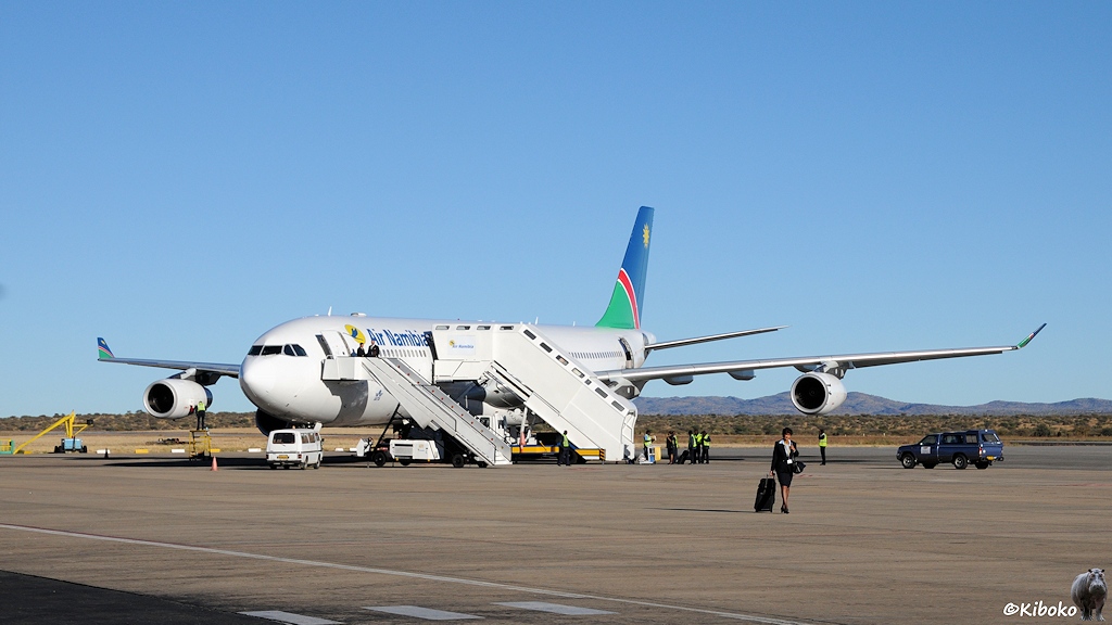 Eine vierstrahlige, weiße Passagiermaschine auf einem Flugfeld mit Ein niedriges Flughafengebäude mit Palmen unter blauem Himmel und strahlender Sonne. Auf dem Rollfeld steht nur eine kleine Probellermaschine.