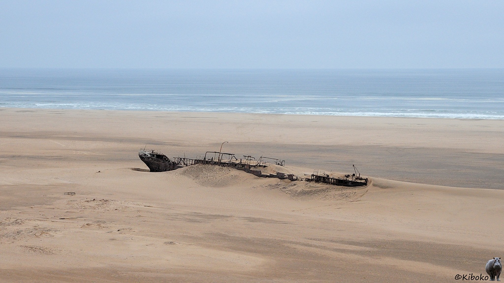 Ein stähllerner Schiffsrumpf liegt in der Wüste. Das Meer ist weiter im Hintergrund zu sehen.