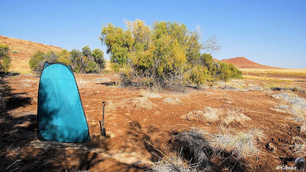 Ein kleines hellblaues Zelt steht auf einer braunen Schotterfläche vor einer weiten Ebene. Vor dem Zelt ist ein Spaten in den Boden gerammt.