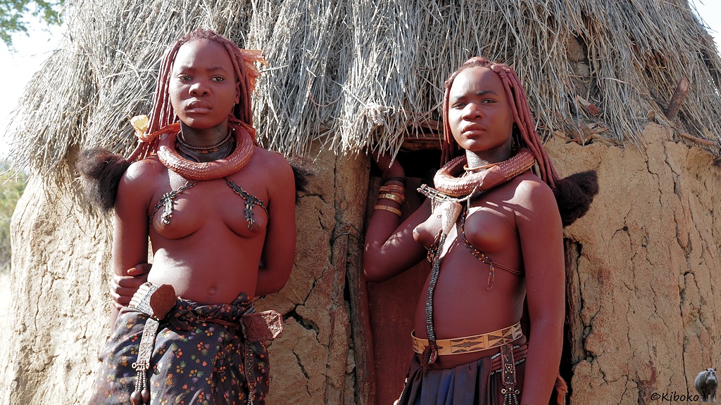 Zwei junge Himbafrauen stehen an der Tür einer strohgedeckten Lehmhütte.