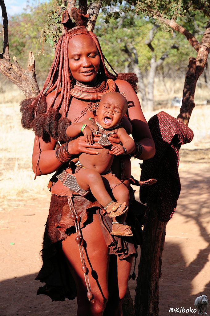 Eine Himbafrau mit langen Zöpfen zeigt Ihren kleinen Jungen. Der schreit laut.