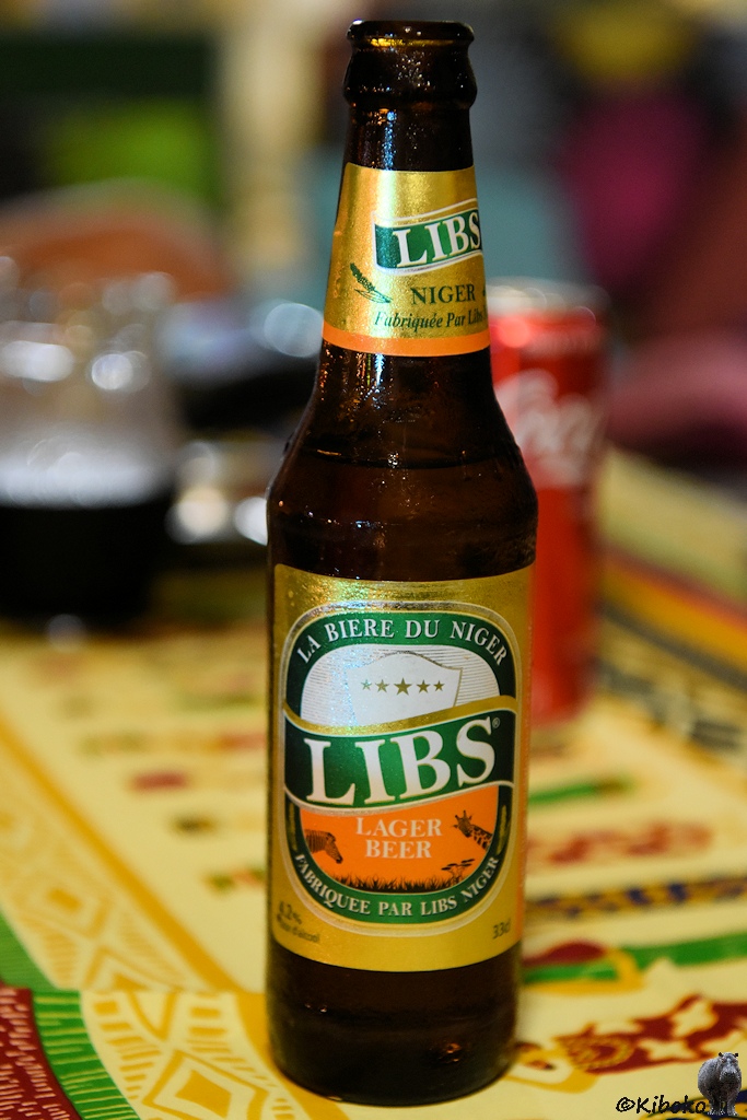 Das Bild zeigt eine Bierflasche auf einer afrikansich gemusterten Tischdecke. Die Bierflasche hat eine Banderole in gold, grün und orange. Die Biermarke ist in großen weißen Buchstaben auf grünem Grund: LIBS.
