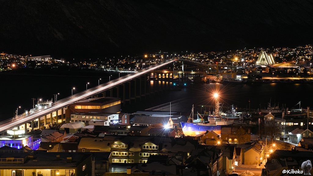 Das Bild zeigt eine nächtlich beleuchtete Straßenbrücke über einen Meeresarm. Im Vordergrund ist eine Werft mit einem blauen Schiff. Im Hintergrund ist eine beleuchtete, dreieckige Kirche.