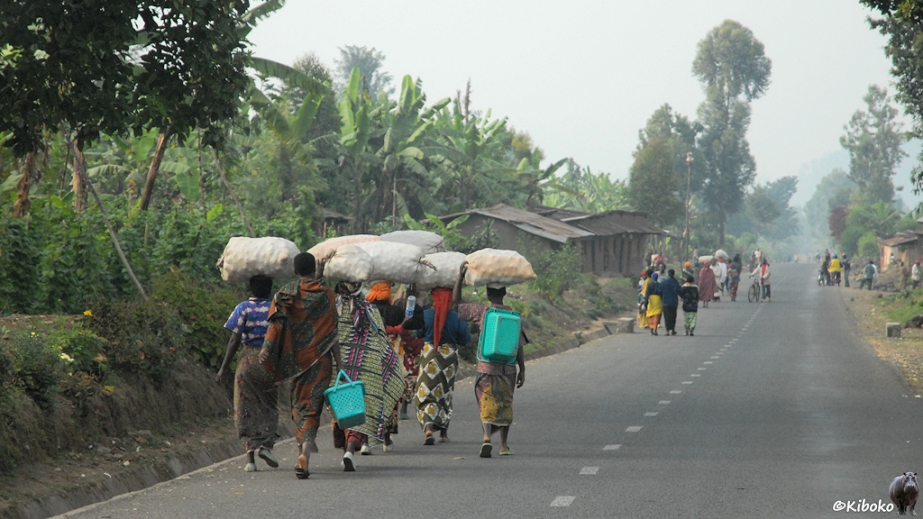 Das Bild zeigt eine gerade Teetrraße mit vielen Menschen auf der Straße. Frauen in bunten Gewändern tragen große, helle Säcke auf den Köpfen.
