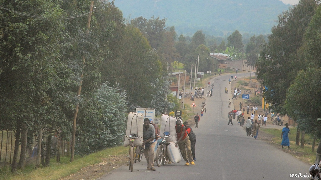 Das Bild zeigt eine abschüssige Teerstraße. Manner schieben Fahrräder die mit großen weißen Säcken beladen sind den Berg hoch. Im Hintergrund laufen viele Menschen auf der Straße.