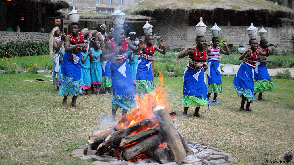 Das Bild zeigt 7 Frauen mit roten Hemden und blauen Röcken beim Tanz am Lagerfeuer. Dabei balancieren sie Körbe auf dem Kopf.