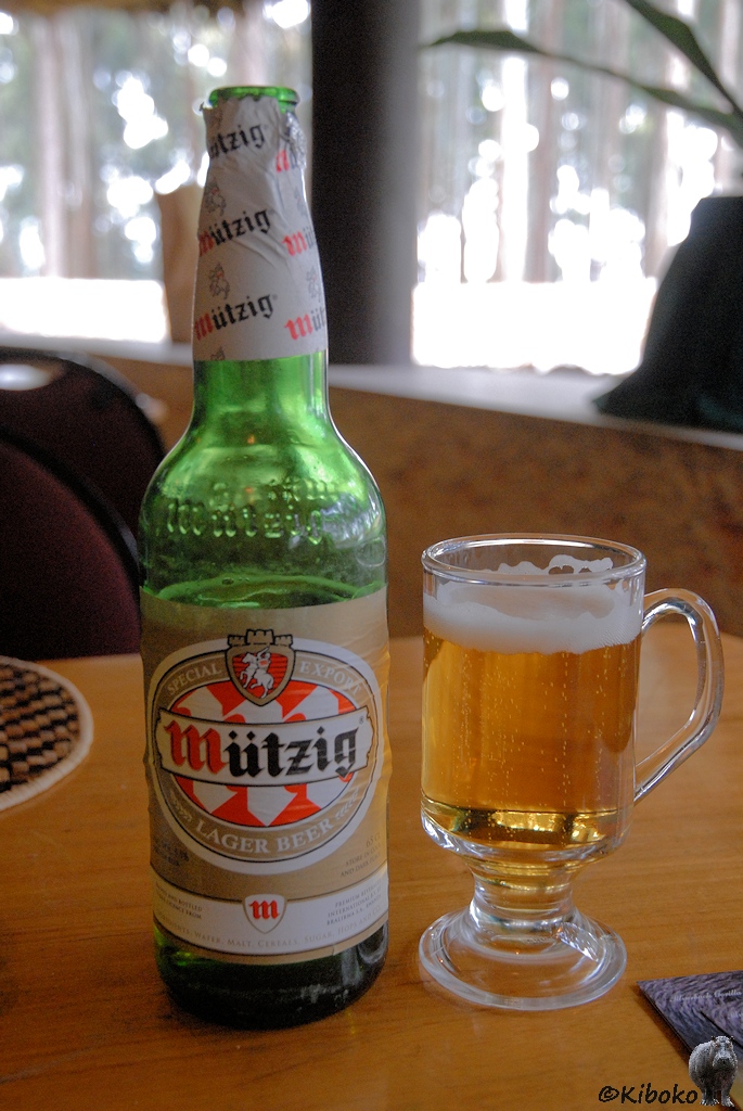Das Bild zeigt eine grüne Bierflasche mit der Aufschrift mit einem roten M und schwarzen ützig. Sowie die Aufschrift Special Export Lager Beer. Daneben steht ein Teeglas mit Henkel, welches mit Bier gefüllt ist.