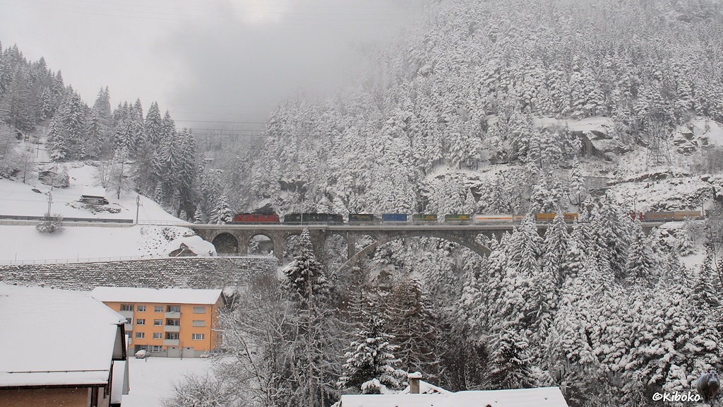 Das Bild zeigt einen Güterzug mit einer roten und einer grünen Elektrolokomotive in einer Winterlandschaft über einer Steinbogenbrücke. Sie ziehen einen Güterzug mit grünen und blauen Wechselpritschen.