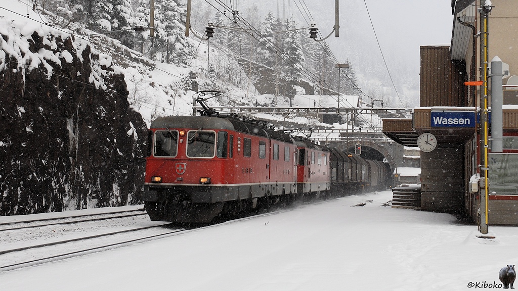 Das Bild zeigt Güterzug mit zwei roten Elektrolokomotiven bei der Fahrt aus einem Tunnel durch einen kleinen Bahnhof im Schneetreiben.