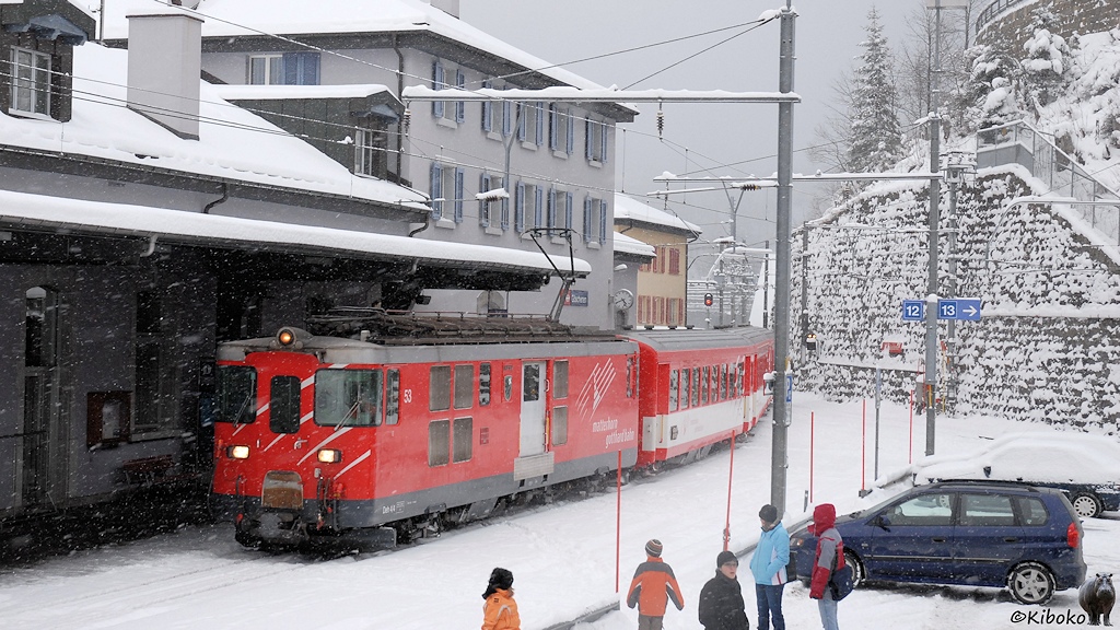 Das Bild zeigt eine rote Schmalspurelektrolok mit grauem Dach und grauem Rahmen. Sie wartet mit einen Personenzug aus rot-weißen Wagen an einem Bahnhofsgebäude im Schneetreiben.