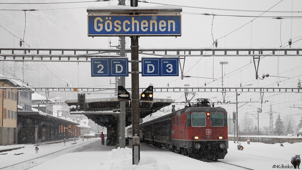 Das Bild zeigt eine rote Elektrolokomotive am verschneiten Mittelbahnsteig. In Bahnsteigmitte steht ein blaues Schild mit der Aufschrift Göschenen.