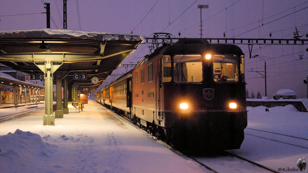Das Bild zeigt einen Personenzug mit einer roten Elektrolokomotive am nächtlich beleuchteten Bahnsteig im Schneetreiben.
